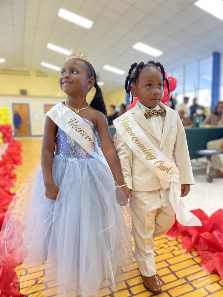 A child in a dress and a child in a suit, both with sashes, walk down a yellow brick road with red poppies.
