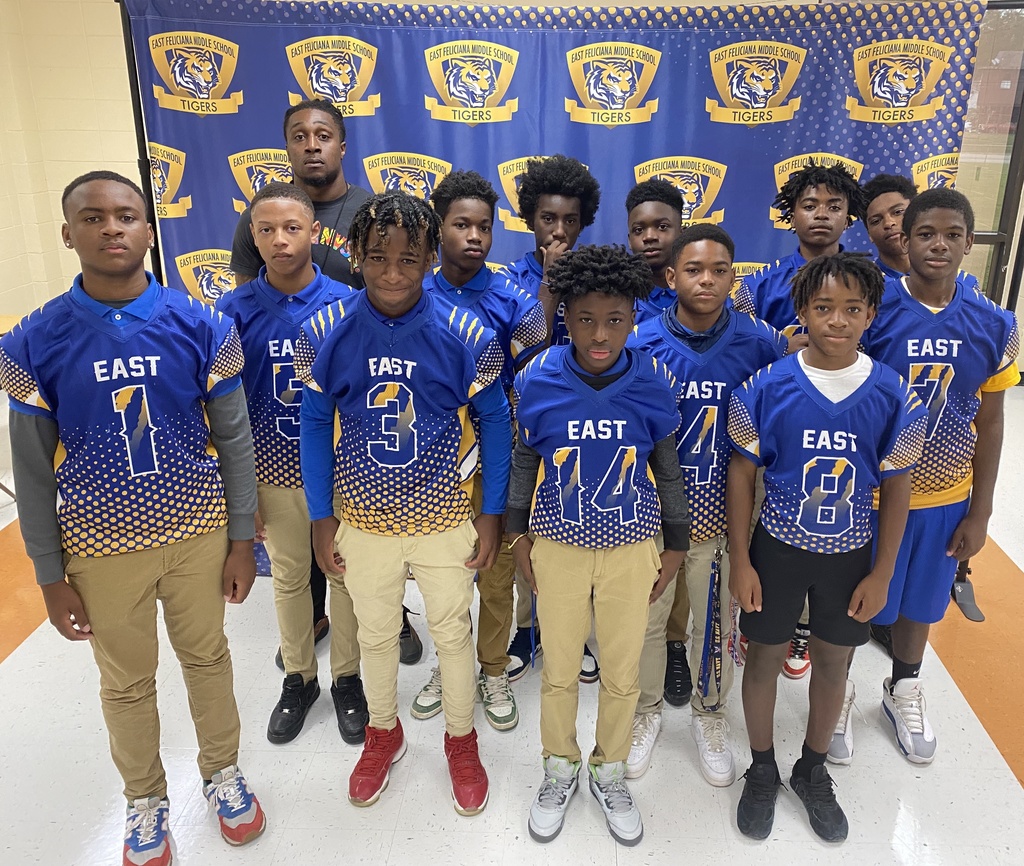 A group of students wearing football uniforms and a coach in front of a blue East Feliciana Middle School backdrop.