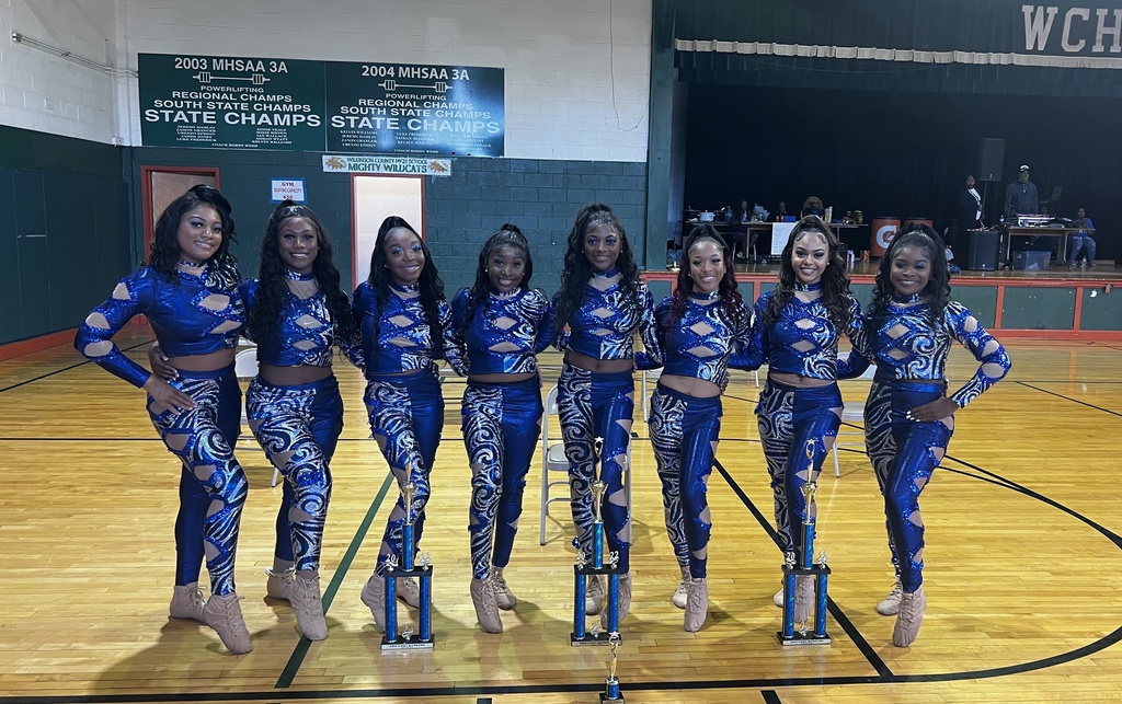 A group of 8 students wearing sparkly outfits stands in a gymnasium. In front of them are several trophies.