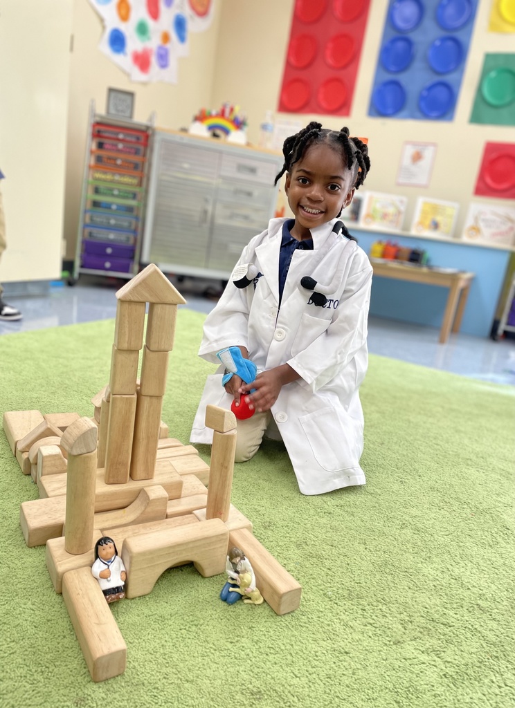 A child is wearing a long white coat. In front of the child are blocks arranged to look like a buildiing.