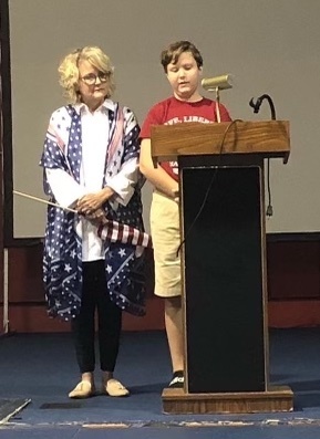 A child is speaking at a podium as an adult with a flag looks on.