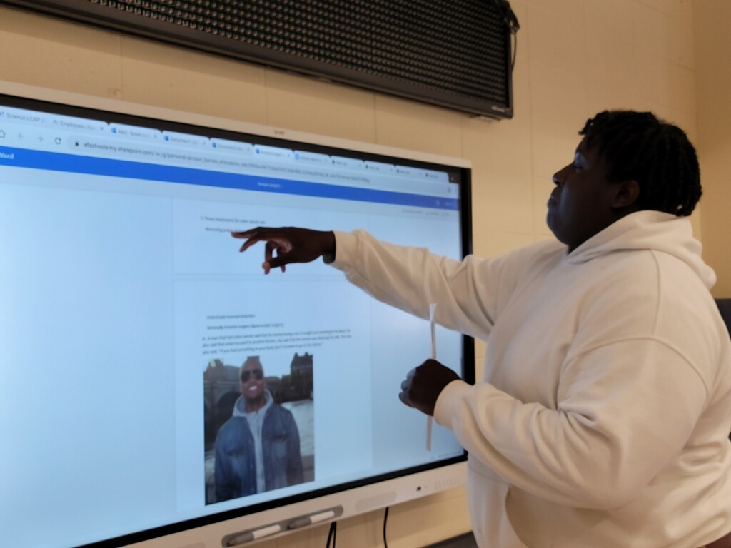 A student touches a smartboard. There is a presentation on the screen.