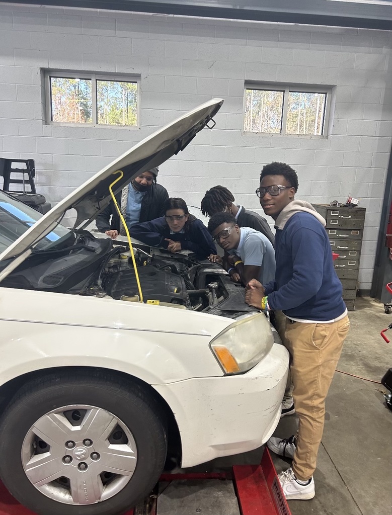 A group of students in safety glasses stand in front of a car with an open hood