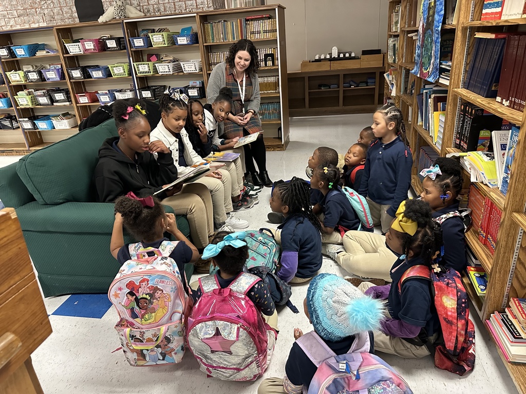 A group of older students sitting on a couch read to a group of younger students sitting in a library as an adult smiles.
