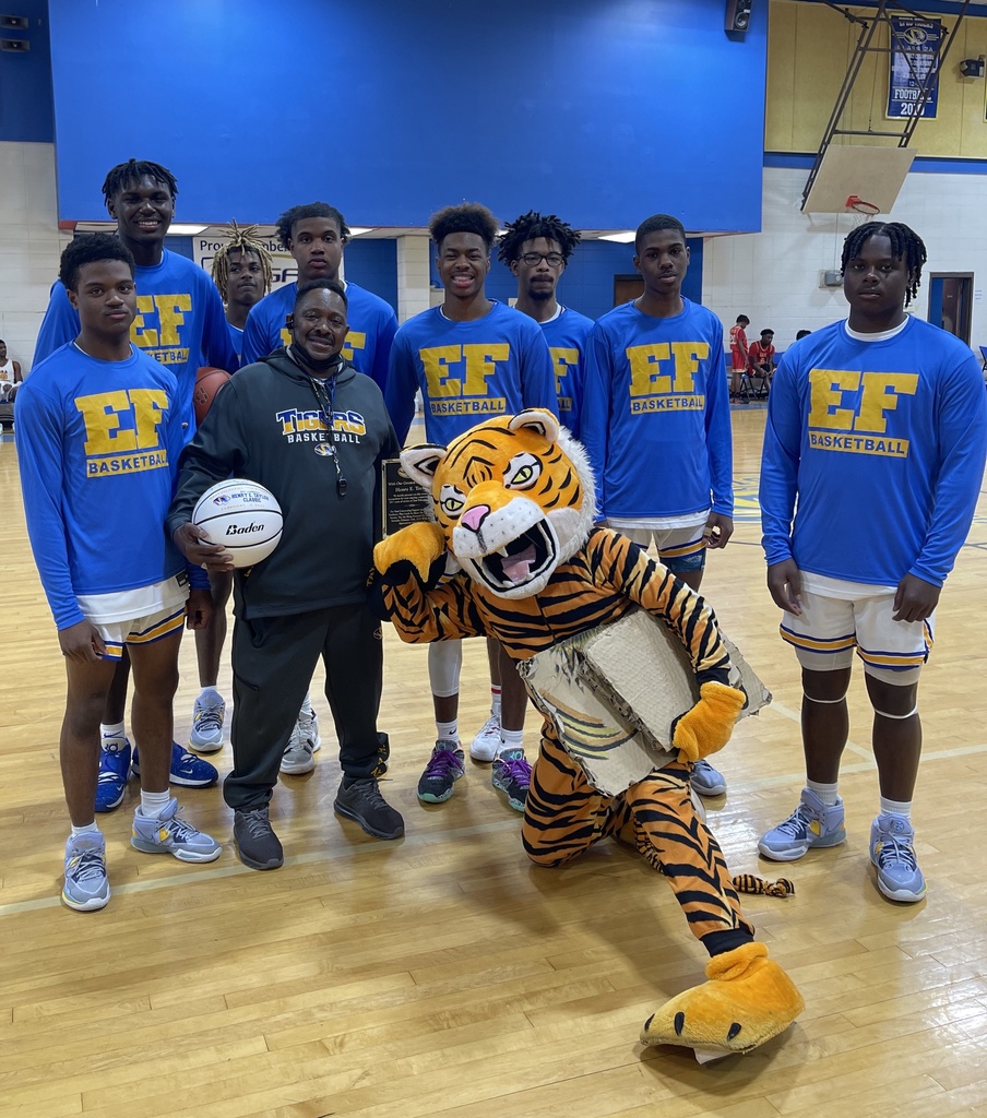 Eight students in blue long-sleeved t-shirts stand with a man holding a signed basketball and a Tiger mascot inside a gymnasium.