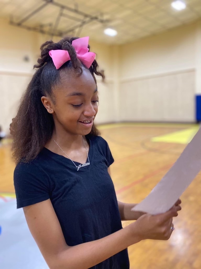 A smiling student in a pink bow looks at a sheet of paper.