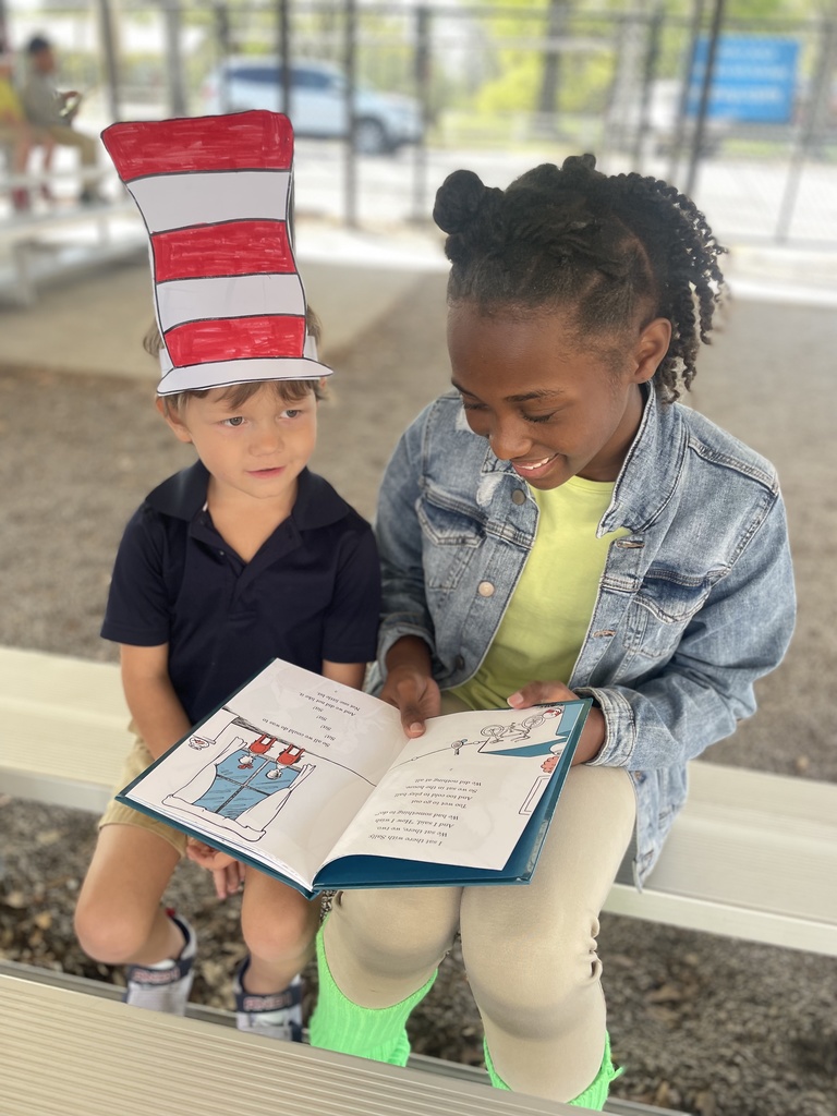 A smiling older student is reading to a younger student in a red and white striped paper hat.