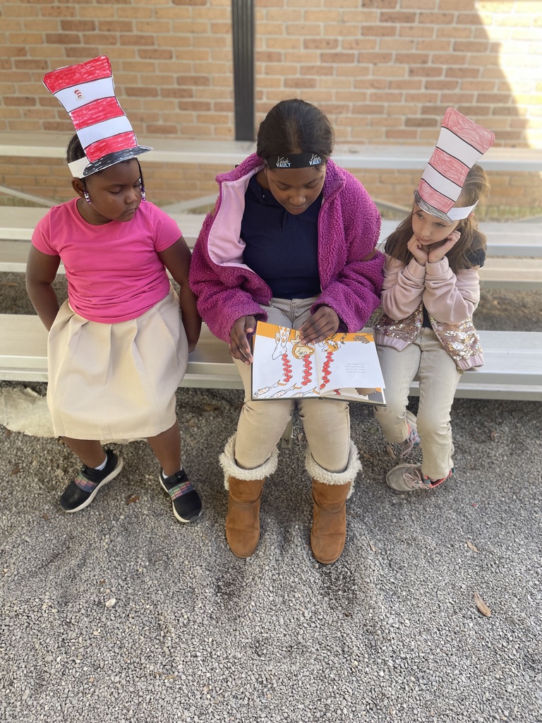 An older student reads to two younger students in red and white striped paper hats