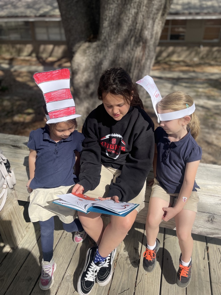 An older student reads to two younger students in red and white striped paper hats.