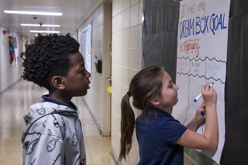 A student writes with a marker on a poster in a hallway as another student looks on.