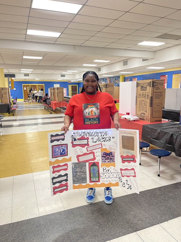 A student in a hall filled with displays holds up a display about Henrietta Lacks.