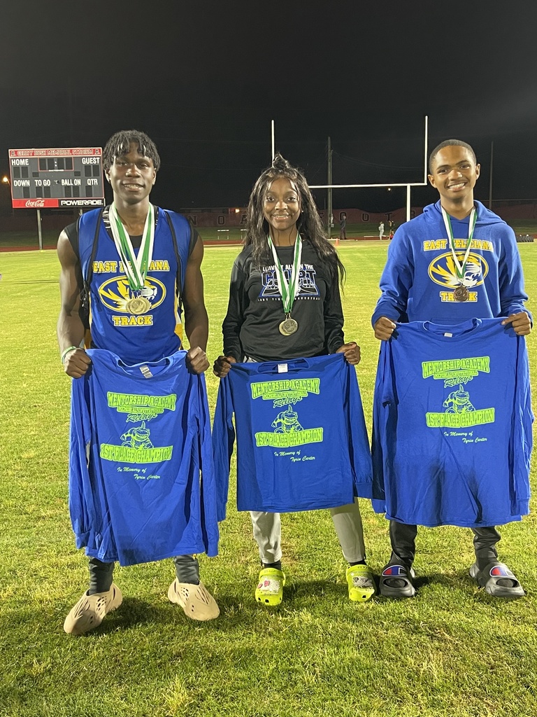 Three students in track gear are standing on a football field. They are also wearing medals and holding blue shirts with neon green text.