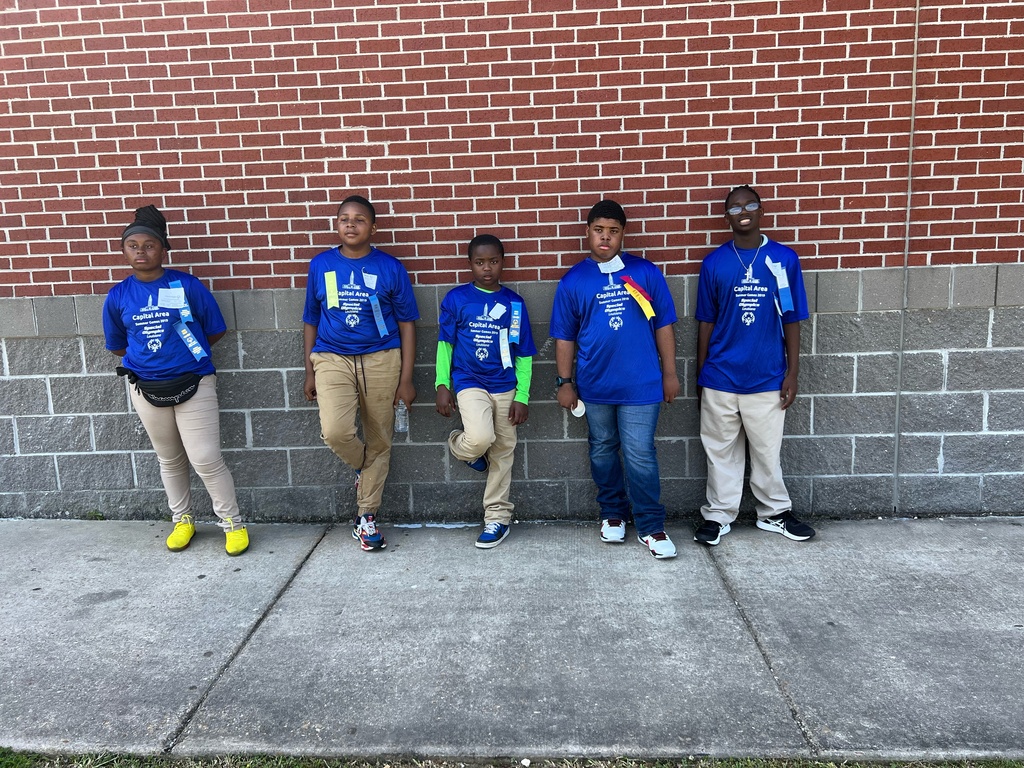 Five students in blue shirts are against a brick backdrop.