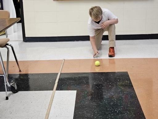 A student rolls a ball next to some rulers on the floor.