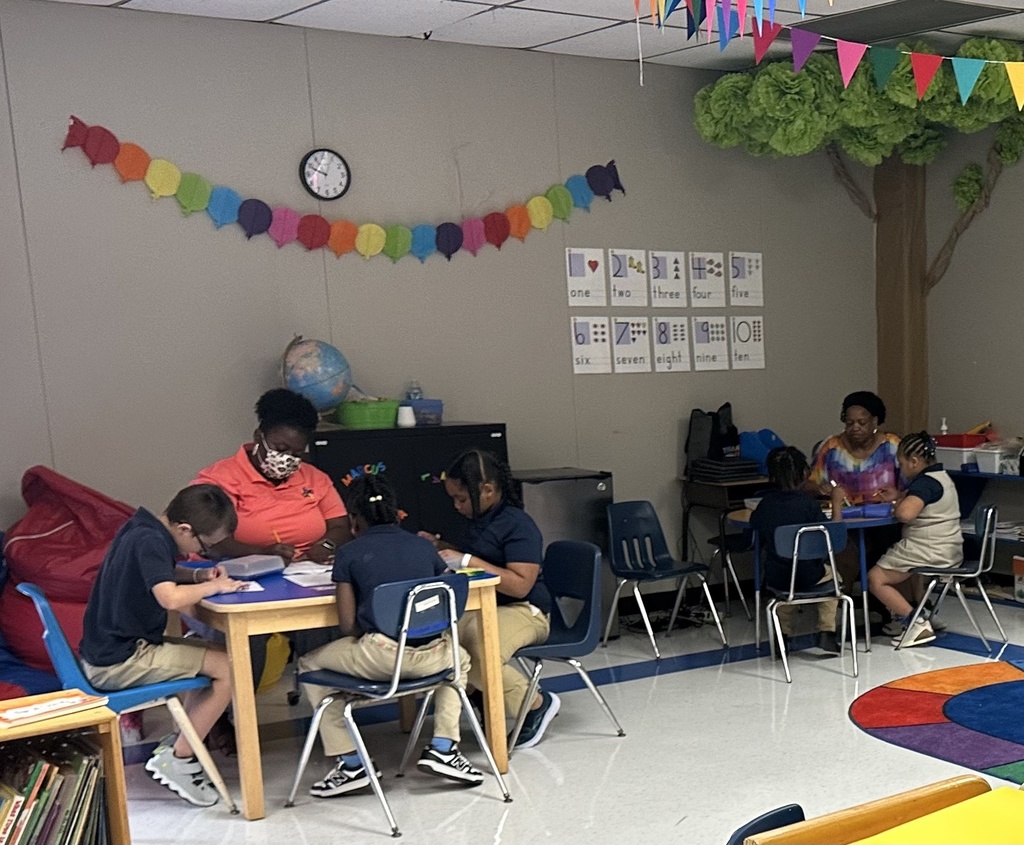 Two teachers sit at tables with small groups of students in a festively decorated classroom.