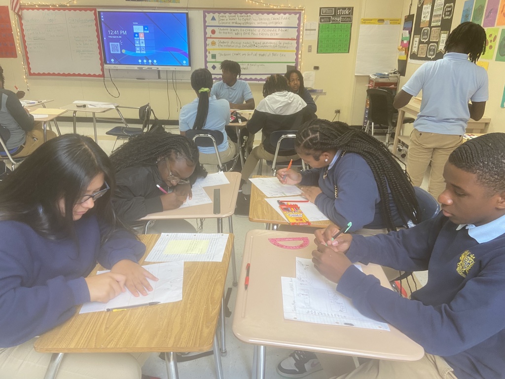 Four students sitting at desks arranged in a group write on pieces of paper in a classroom.